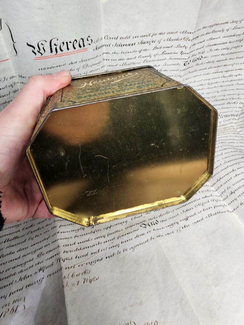 May include: An antique, octagonal tin with a gold interior and a green and gold exterior. The tin is held over a document with handwritten text. The word "Hereas" is visible on the document.