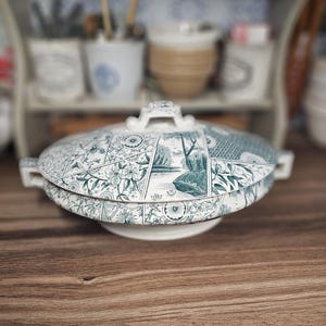 May include: A white and teal ceramic serving dish with a lid. The lid features a detailed floral and landscape design. The dish has handles on either side and a small handle on top of the lid. The dish is sitting on a wooden surface.