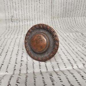 May include: A circular, antique metal object with a copper centre and a decorative, textured border. The outer ring has a bronze-like finish, and the object rests on a document with handwritten script. The item is likely vintage.