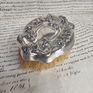 Puede incluir: Cepillo antiguo plateado con detalles florales y de volutas ornamentados. El cepillo tiene forma rectangular con una parte superior curvada y una base de cerdas naturales. El cepillo está sobre un documento manuscrito.