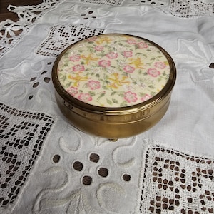 Könnte beinhalten: Eine runde, messingfarbene Schmuckdose mit floralem Design auf dem Deckel. Der Deckel hat einen cremefarbenen Hintergrund mit rosa Blüten und gelben Akzenten. Die Dose liegt auf einer weißen, bestickten Tischdecke.