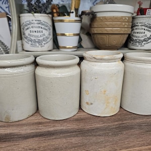 May include: Four cylindrical stoneware jars with a smooth, off-white finish and a distinct rim at the top. Each jar features a simple, utilitarian design with subtle variations in texture and color, including some light brown staining on one jar.