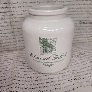 Pode incluir: Pote de cerâmica branco com formato cilíndrico. O pote apresenta um rótulo verde e branco com o texto "Edmond Fallot" e uma ilustração de um edifício. O pote é provavelmente para armazenar alimentos.