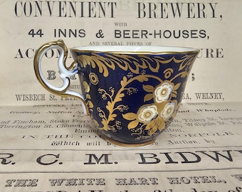 May include: A navy blue teacup with gold floral designs and a white handle. The teacup has a gold rim and a white floral design. The handle is white with gold accents. The teacup is on a vintage newspaper background.