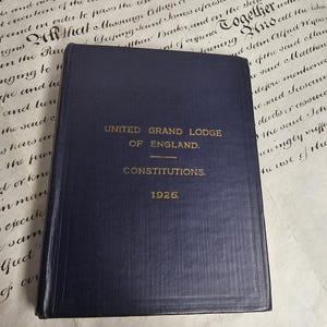 Peut inclure: Un livre bleu marine intitulé "UNITED GRAND LODGE OF ENGLAND. CONSTITUTIONS. 1926." Le livre est posé sur un fond de parchemin vieilli avec une écriture manuscrite.