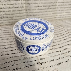 Può includere: Una latta di Borax vintage, rotonda, con un design blu e bianco. Il coperchio presenta il testo "THE PATENT BORAX CO LTD" e "BIRMINGHAM AND LONDON". La latta è posta su uno sfondo di carta invecchiata con scritte a mano.