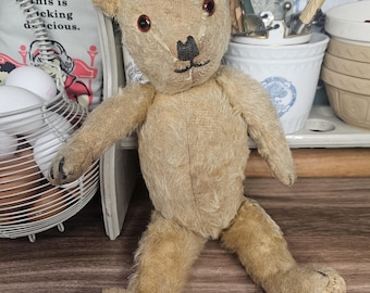 May include: A vintage teddy bear with light brown fur, a black stitched nose, and dark red eyes. The bear is sitting with its arms and legs spread. The bear's fur shows signs of wear and age.
