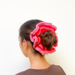 May include: A handmade, crochet scrunchie in shades of red and pink. The scrunchie is worn in the hair, with the bun visible in the center. The crochet design creates a ruffled, flower-like appearance, adding a pop of color.