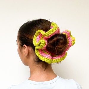 May include: A large, handmade crochet hair accessory in a flower shape. The accessory features alternating stripes of bright pink and lime green. The hair piece is worn in a bun.