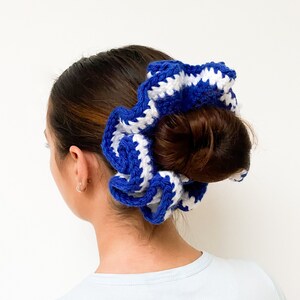 May include: A blue and white crocheted scrunchie worn in a bun. The scrunchie is made of thick yarn and has a wavy, textured appearance. The color scheme is a bold blue and white, creating a striking contrast. The hair accessory adds a touch of handmade charm.