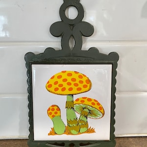 May include: Green cast iron hot pad with a white ceramic tile featuring a yellow and orange mushroom with white polka dots.