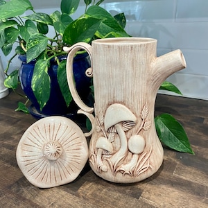 Può includere: Una teiera in ceramica con un design a forma di fungo. La teiera è beige con accenti marroni e ha un manico e un beccuccio. La teiera è appoggiata su una superficie di legno.