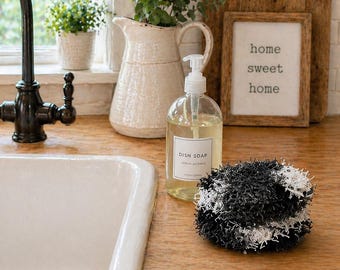 May include: A kitchen scene with a white sink, black faucet, and a clear dish soap bottle. A black and white scrubby sits on the wooden countertop. A sign that says "home sweet home" is in the background.