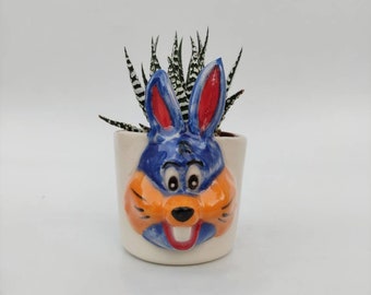 May include: A white ceramic planter shaped like a cartoon bunny face with blue ears, orange fur, and a red mouth. A small succulent plant with green and white striped leaves is growing in the planter.