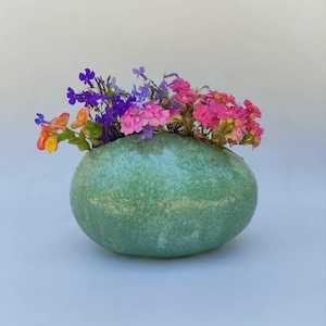 May include: A green ceramic vase shaped like an egg with a speckled texture, filled with a bouquet of purple, pink, and orange flowers.