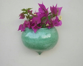 May include: A green ceramic planter with a speckled finish hangs on a white wall. It is filled with pink bougainvillea flowers.
