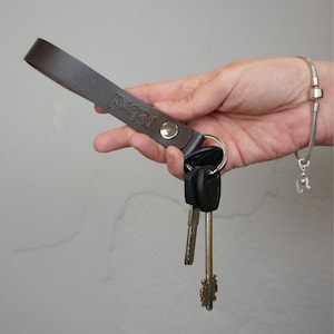 May include: A brown leather keychain with the word "Relard" embossed on the strap. The keychain is attached to a silver keyring holding several keys. A silver bracelet is visible in the background.