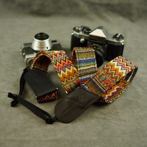May include: A woven camera strap featuring a multi-colored zigzag and geometric pattern in shades of red, blue, yellow, and green. The strap includes black leather end tabs embossed with the brand name Velard and black nylon adjustment webbing.