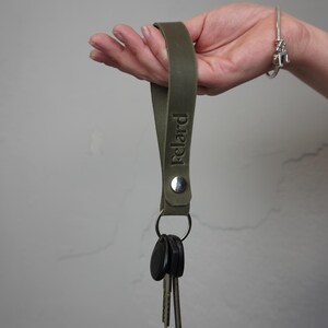 Puede incluir: Llavero de cuero verde oliva con el nombre "Felard" grabado en relieve. El llavero está sujeto a un anillo de metal plateado que sujeta varias llaves. Un broche de metal plateado asegura el lazo de cuero.