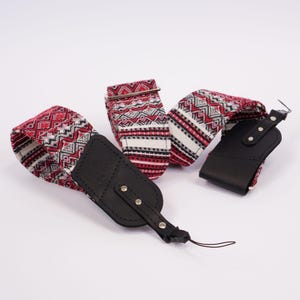 May include: A camera strap featuring a woven fabric pattern in red, white, and black with geometric diamond and stripe motifs. The strap includes black leather end tabs with silver-toned metal rivets and a thin black cord attachment loop.
