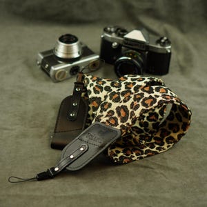 May include: A camera strap featuring a beige and black leopard print fabric pattern with black leather end tabs. The leather tabs are secured with silver-toned metal rivets and feature the embossed brand name Relard on the front surface.
