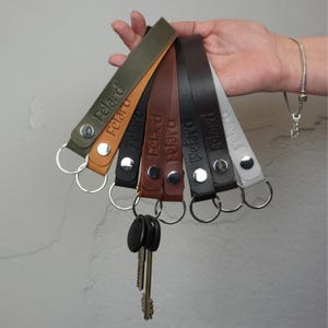 May include: A hand displays six leather keychains in various colors: olive green, tan, black, brown, dark grey, and white. Each keychain has a silver ring and a snap closure. The word "Felard" is embossed on each strap.