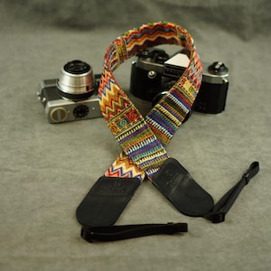 May include: A camera strap featuring a vibrant, multi-coloured woven pattern with zig-zag and geometric motifs. The strap includes black leather end tabs embossed with the brand name Velord and adjustable black nylon webbing connectors for attachment.