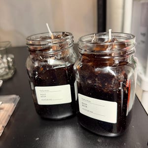 May include: Two glass jar candles filled with dark brown gel wax, resembling root beer. Each candle has a white label and a single white wick. The jars sit on a dark surface.