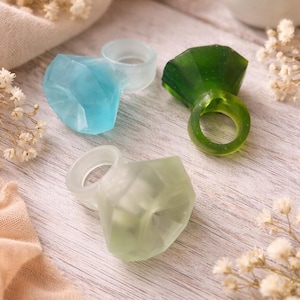 May include: Four colorful resin rings in shades of blue, green, and clear, each with a faceted gem-like design. The rings are displayed on a light wooden surface, with a candle and floral accents.