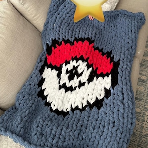 May include: A blue knitted blanket with a large red, white, and black Poké Ball design. The blanket is made of chunky yarn and has a soft, fuzzy texture.