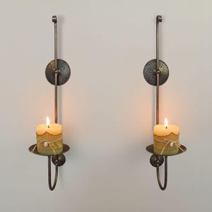 Puede incluir: Dos candelabros de pared con velas encendidas. Los soportes son de metal oscuro con una placa trasera circular y un brazo largo y delgado. Cada vela es amarilla y está envuelta con hilo decorativo.