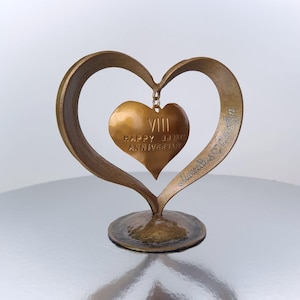 Puede incluir: Una escultura en forma de coraz&oacute;n de color bronce con un colgante de coraz&oacute;n m&aacute;s peque&ntilde;o. El colgante dice "VIII HAPPY BRONZE ANNIVERSARY". La escultura tiene una base y est&aacute; firmada "Marcella's Crafts".