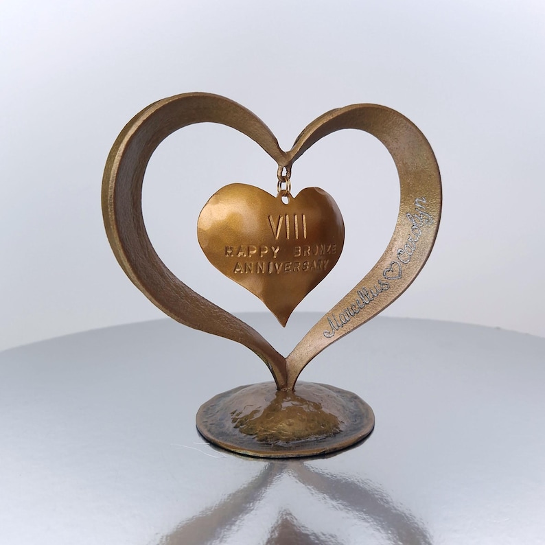 Puede incluir: Una escultura en forma de coraz&oacute;n de color bronce con un colgante de coraz&oacute;n m&aacute;s peque&ntilde;o. El colgante dice "VIII HAPPY BRONZE ANNIVERSARY". La escultura tiene una base y est&aacute; firmada "Marcella's Crafts".