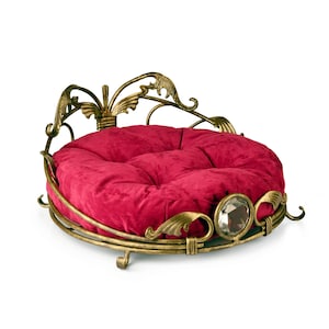 Puede incluir: Una cama para perros de terciopelo rojo con un marco de metal dorado. El marco es ornamentado y presenta un diseño decorativo. La cama es redonda y tiene un cojín en el centro.