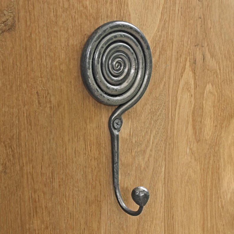 May include: A dark gray metal wall hook featuring a circular spiral design at the top and a curved hook extending downward. The metal has a textured, hammered finish and is secured to the surface with a single visible screw in the center.