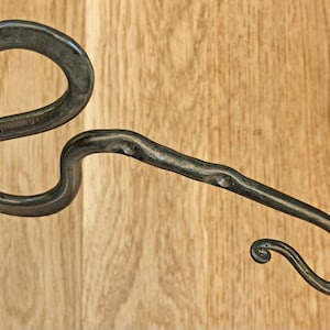 Pu&ograve; includere: Un gancio da parete in metallo forgiato a mano di colore scuro, caratterizzato da un design curvo e sinuoso con una superficie ruvida e irregolare. Presenta un'ampia asola sul lato sinistro e un braccio allungato che termina con un ricciolo.