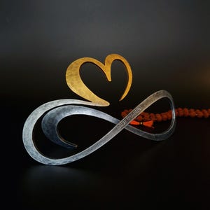 Op de afbeelding: Een metalen sculptuur met een zilverkleurig oneindigheidssymbool verweven met een goudkleurig hart. Het zilverkleurige deel heeft een gehamerde textuur en is gegraveerd met de tekst DAVE & SHEILA. Het object bestaat uit twee metalen elementen.