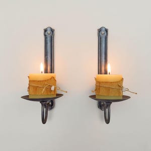 May include: Two wall-mounted candle holders with lit square candles. The candles are wrapped in a textured material and tied with twine. The holders are dark metal with a decorative design and a curved handle.