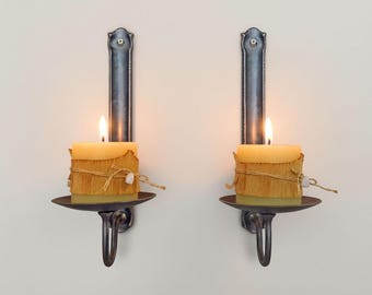 May include: Two wall-mounted candle holders with lit square candles. The candles are wrapped in a textured material and tied with twine. The holders are dark metal with a decorative design and a curved handle.