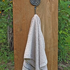 May include: A dark gray metal hook with a circular spiral design at the top, mounted on a vertical wooden board. A light beige terry cloth towel with a thin dark horizontal stripe near the bottom edge hangs from the hook.
