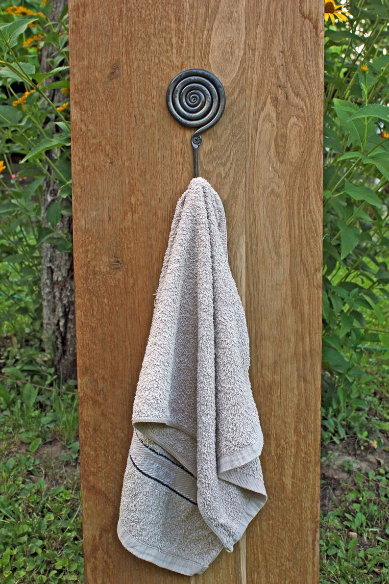 May include: A dark gray metal hook with a circular spiral design at the top, mounted on a vertical wooden board. A light beige terry cloth towel with a thin dark horizontal stripe near the bottom edge hangs from the hook.