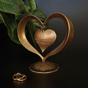 Puede incluir: Una escultura en forma de coraz&oacute;n de color bronce con un colgante de coraz&oacute;n m&aacute;s peque&ntilde;o con la inscripci&oacute;n "VIII" y "BRONZE ANNIVERSARY". La escultura tiene una base texturizada y la fecha "05-05-18" est&aacute; grabada en el lateral. Dos anillos de oro en la base.