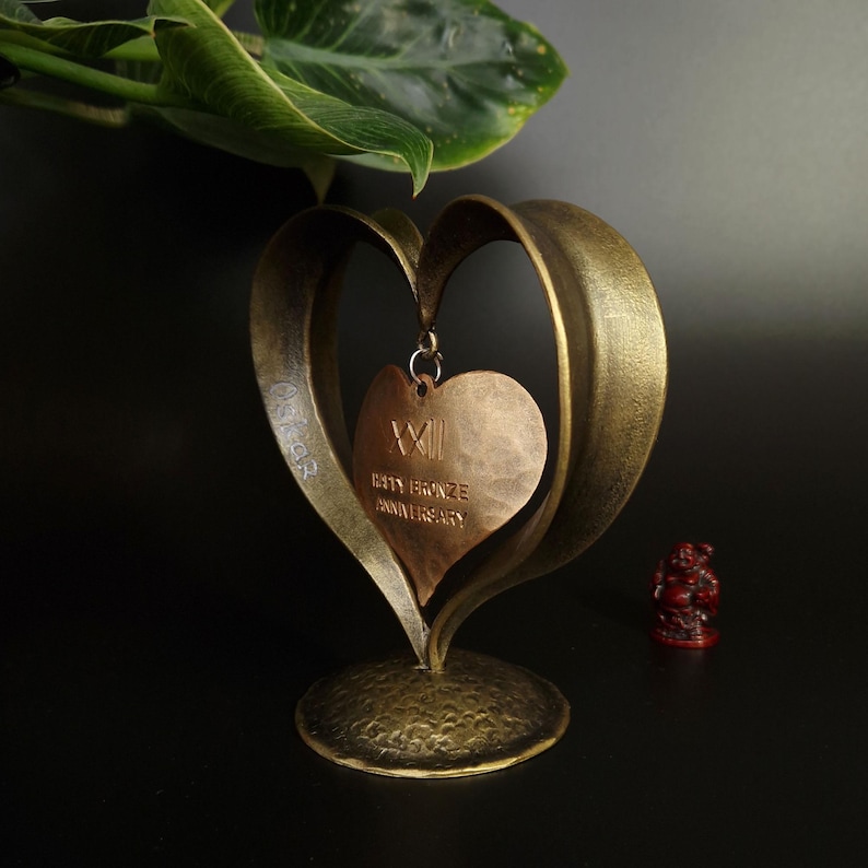 Puede incluir: Escultura en forma de coraz&oacute;n de color bronce con un colgante que dice "XXII" y "BRONZE ANNIVERSARY". La escultura tiene un acabado texturizado y se asienta sobre una base redonda. Una peque&ntilde;a figura roja est&aacute; en el fondo.