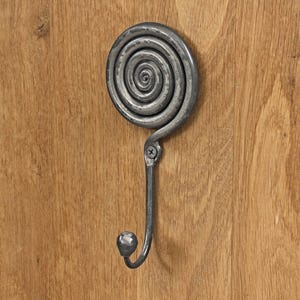 May include: A dark gray metal wall hook featuring a circular spiral design at the top and a curved hook ending in a small rounded knob. The metal has a textured, hammered finish and is secured to a light brown wooden surface with a single screw.