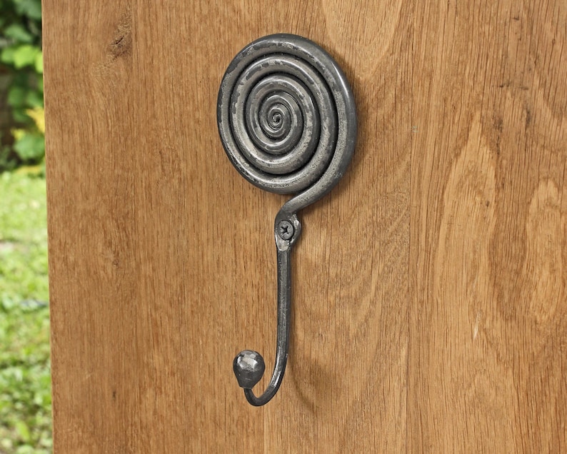 May include: A dark gray metal wall hook featuring a circular spiral design at the top and a curved hook ending in a small rounded knob. The metal has a textured, hammered finish and is secured to a light brown wooden surface with a single screw.
