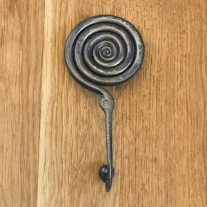 May include: A dark gray metal wall hook featuring a prominent circular spiral design at the top. The hook extends downward from the center of the spiral and terminates in a small rounded knob at the bottom. A single mounting hole is visible.