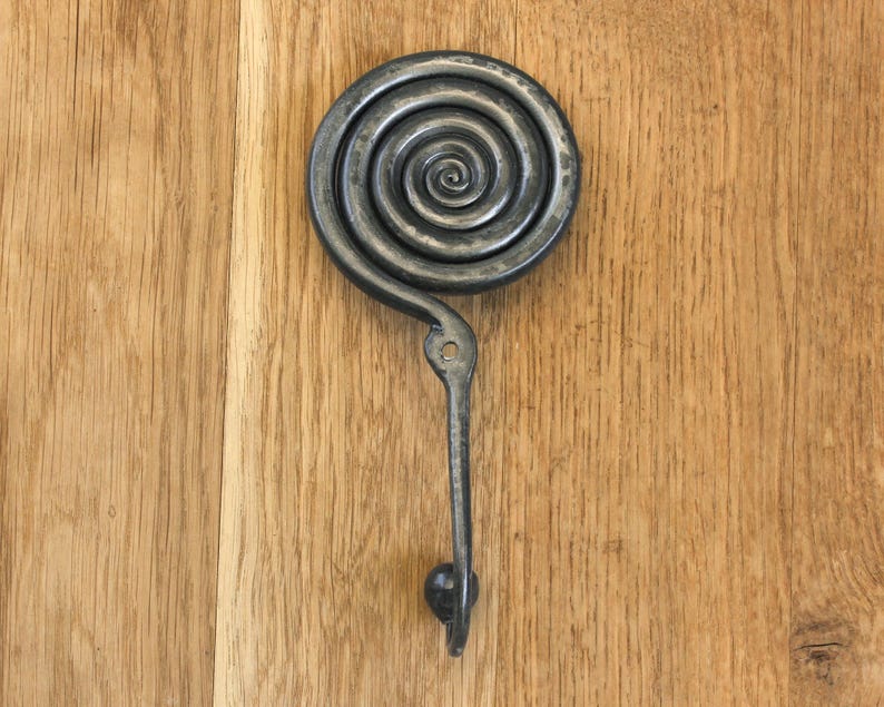 May include: A dark gray metal wall hook featuring a prominent circular spiral design at the top. The hook extends downward from the center of the spiral and terminates in a small rounded knob at the bottom. A single mounting hole is visible.