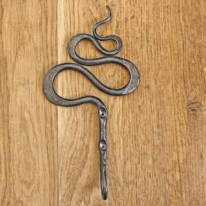 Pu&ograve; includere: Un gancio da parete in metallo grigio scuro forgiato a mano, caratterizzato da un design sinuoso a forma di serpente nella parte superiore e da uno stelo verticale dritto con due fori circolari preforati per le viti, con finitura martellata.