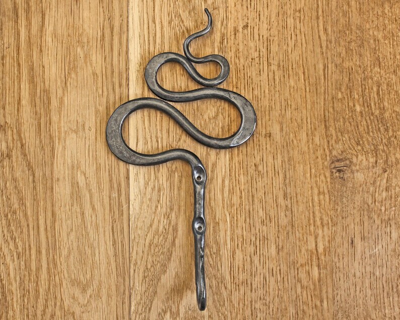 Pu&ograve; includere: Un gancio da parete in metallo grigio scuro forgiato a mano, caratterizzato da un design sinuoso a forma di serpente nella parte superiore e da uno stelo verticale dritto con due fori circolari preforati per le viti, con finitura martellata.