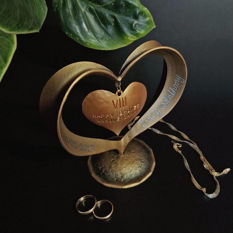 Puede incluir: Una escultura de aniversario en forma de coraz&oacute;n de color bronce con la inscripci&oacute;n "Happy Bronze Anniversary" y el n&uacute;mero romano VIII. La escultura tiene una base texturizada e incluye los nombres "Emily Anthony" y la fecha "10-02-2017". Dos anillos de oro en la parte inferior.